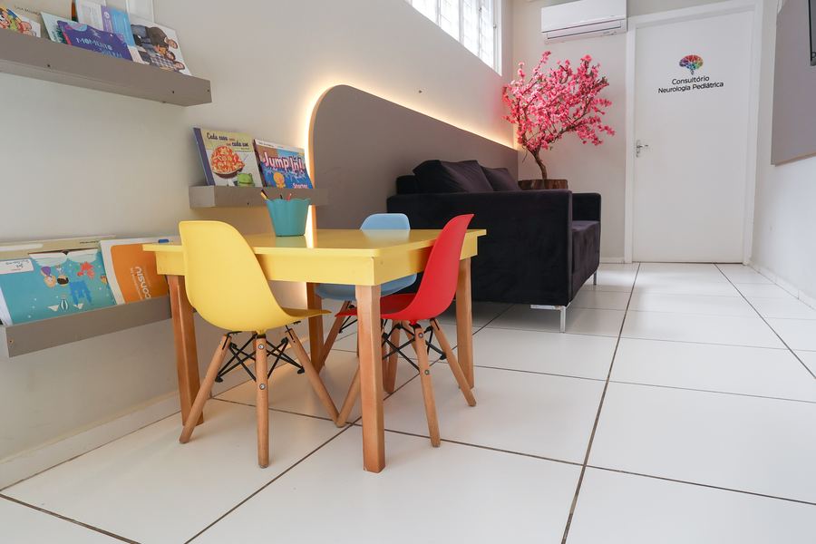 Sala de espera reservada com sofá escuro, mesa infantil amarela e prateleiras de livros