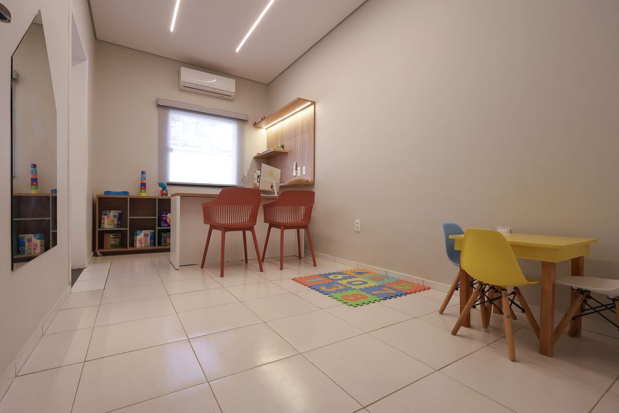 Sala de Fonoaudiologia com mesa infantil amarela, tapete e prateleiras de jogos